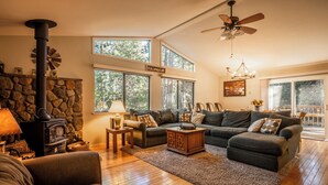 A cozy living room with a stone fireplace, large windows, and a ceiling fan. - Bennetts’ Bear Den | Game Room • Cozy Forest Cabin (Dorrington)