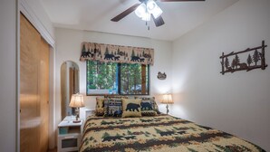 A bedroom with a bed, two bedside lamps, a window with a forest-themed valance, a ceiling fan, and a wall-mounted decoration. - Bennetts’ Bear Den | Game Room • Cozy Forest Cabin (Dorrington)