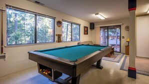 A pool table in a room with large windows, a door leading outside, and a small shelf with items. - Bennetts’ Bear Den | Game Room • Cozy Forest Cabin (Dorrington)