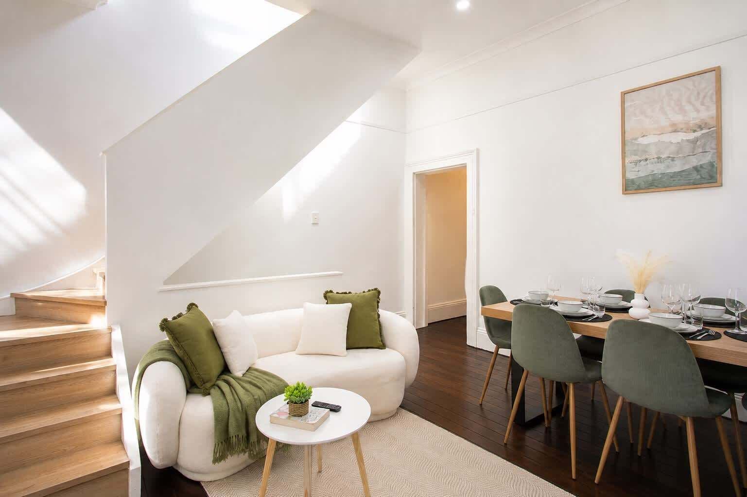 A bright, high-ceilinged living space with a plush white sofa and soft green accents, opening onto a stylish dining area - the perfect spot to unwind or gather over a meal in natural light.