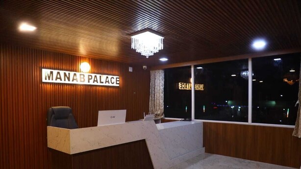 Een receptiebalie met een bord waarop 'MANAB PALACE' staat, en een uitzicht op de stad 's nachts.