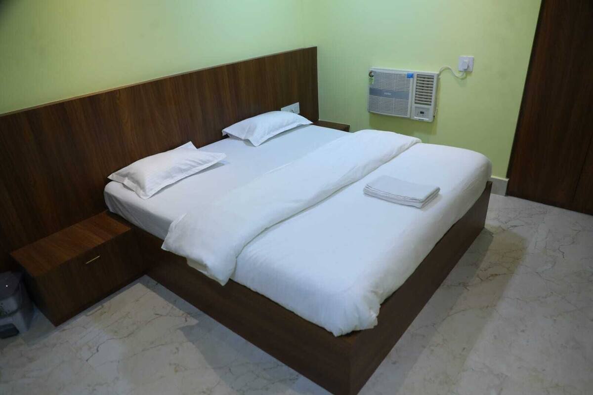 Un lit bien fait, avec du linge de lit blanc et des oreillers, une tête de lit en bois, une petite table de chevet et un appareil de climatisation fixé au mur.