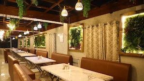 Een restaurant met zitbankjes en tafels, hangende lampen en groene planten aan de muren.