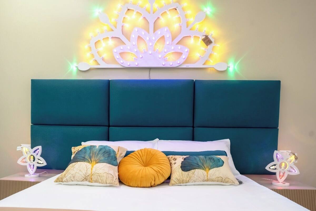 Una cama con cabecera tapizada en color verde azulado, varios almohadones e iluminación decorativa en la pared.