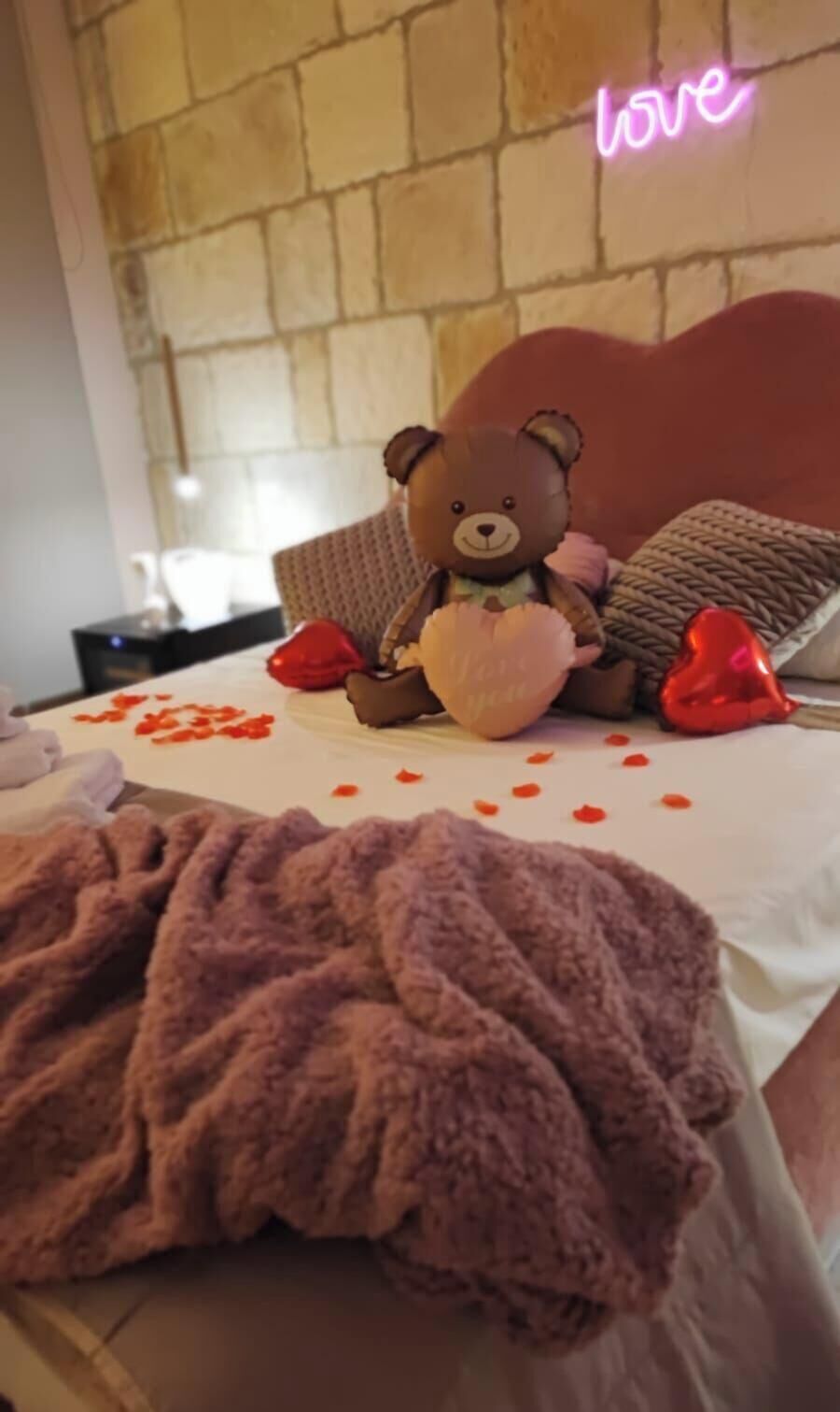 A bed with a teddy bear, a heart-shaped pillow, and scattered rose petals.
