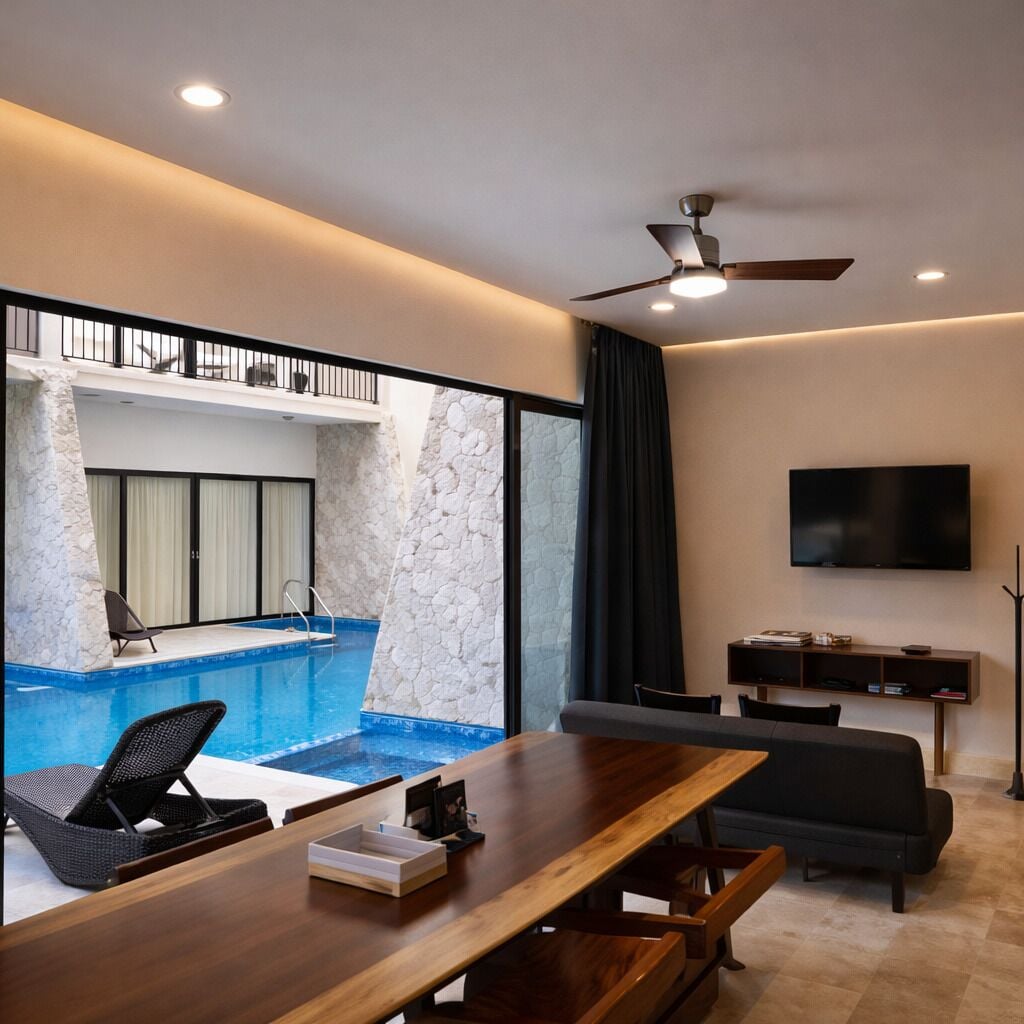 Un salon moderne avec une table à manger en bois, un canapé, un téléviseur fixé au mur et une vue sur une piscine intérieure.