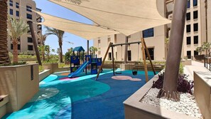 A children's play area with a slide, swings, and a climbing structure, surrounded by palm trees and a pool. - Stayshort - Bright 1 Bedroom in Madinat Jumeirah Lving That Sleeps 2 (Dubai)