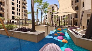 A pool area with a colorful, pebble-lined pathway, surrounded by buildings with balconies and a clear sky. - Stayshort - Bright 1 Bedroom in Madinat Jumeirah Lving That Sleeps 2 (Dubai)