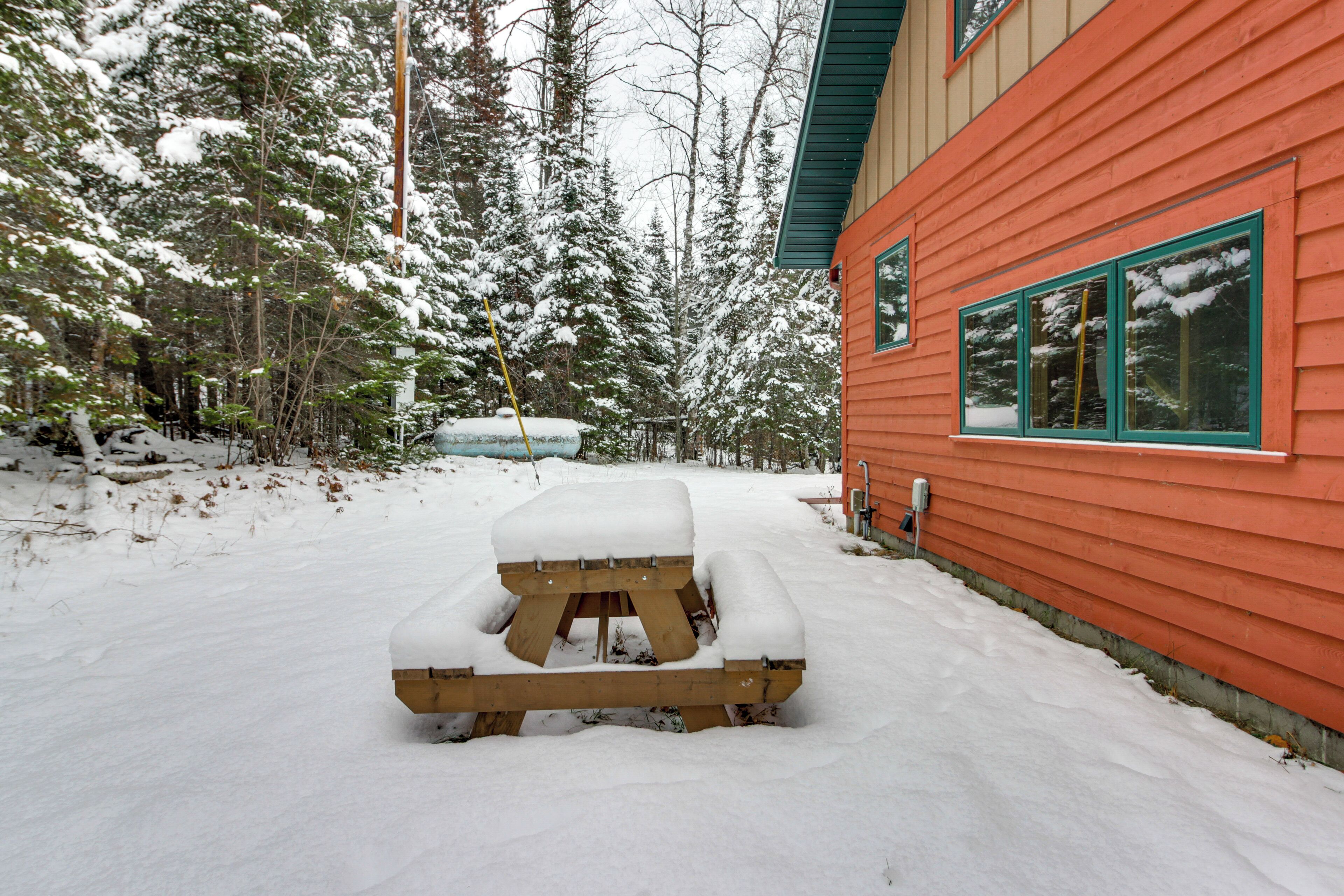 A wooden picnic table covered in snow, a red building with green windows, and a snowy forest in the background.