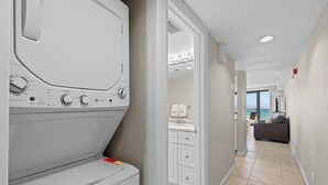A laundry area with a front-loading washer and dryer, a kitchen with white cabinets, and a view of the ocean through a large window. - New! Beachfront Studio with Covered Balcony, Beach Views, Pool, Tram (Miramar Beach)