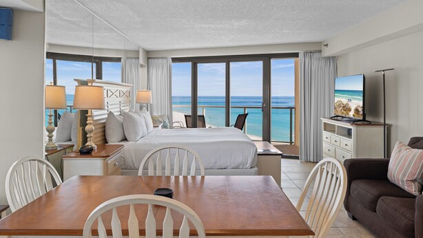 A hotel room with a large bed, a dining table, a TV, and a view of the ocean. - New! Beachfront Studio with Covered Balcony, Beach Views, Pool, Tram (Miramar Beach)