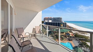 A balcony with chairs, a pool, and a view of the beach and hotel buildings. - New! Beachfront Studio with Covered Balcony, Beach Views, Pool, Tram (Miramar Beach)