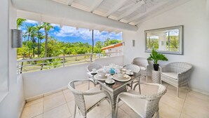 A balcony with wicker furniture, a dining table set with plates and cups, and a view of palm trees and a clear sky. - Glitter Bay Estate 414 1 Bedroom Apts (Holetown)