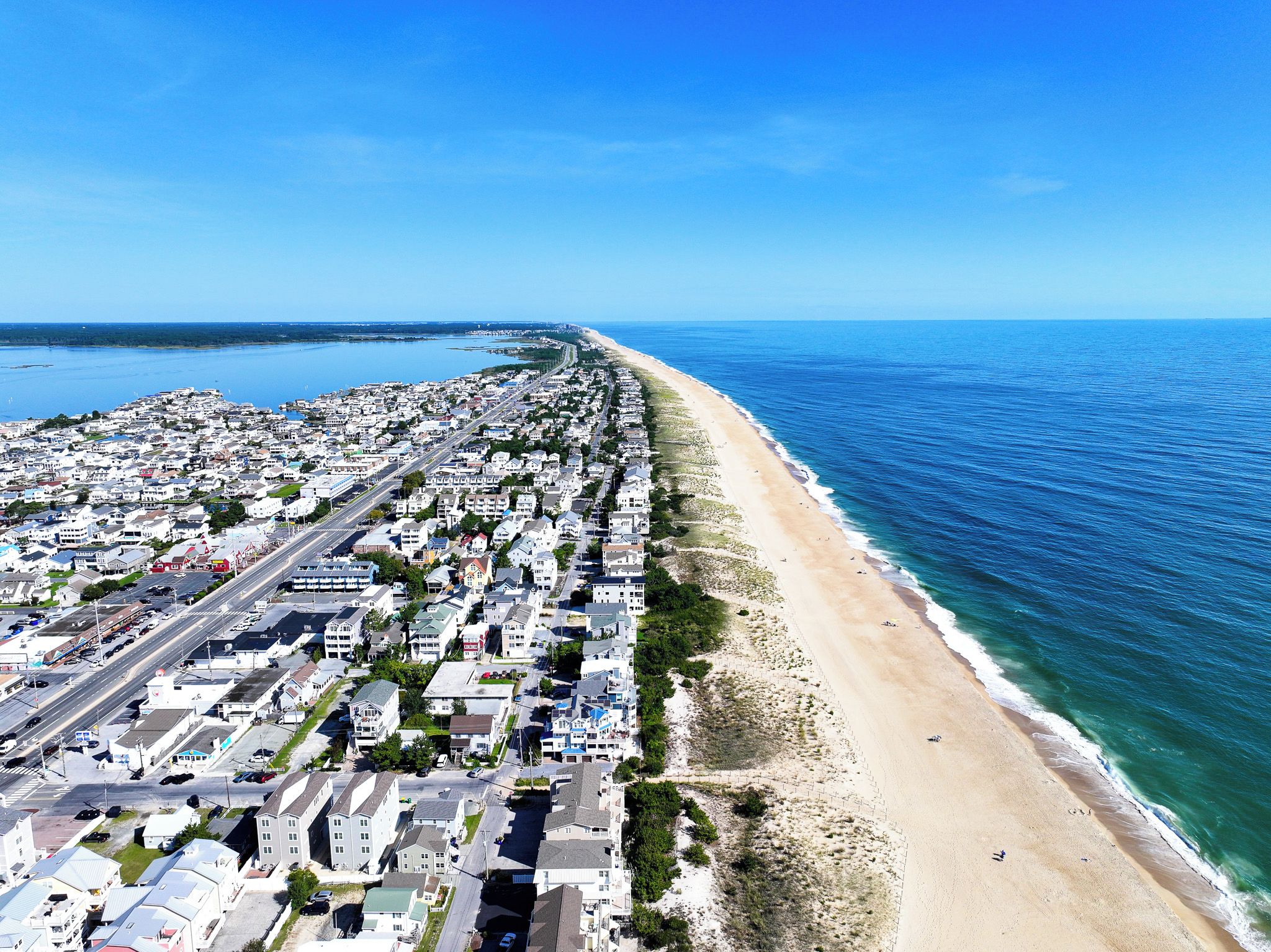 An aerial view of a coastal town with a long beachfront, dense residential area, and a clear blue sky.