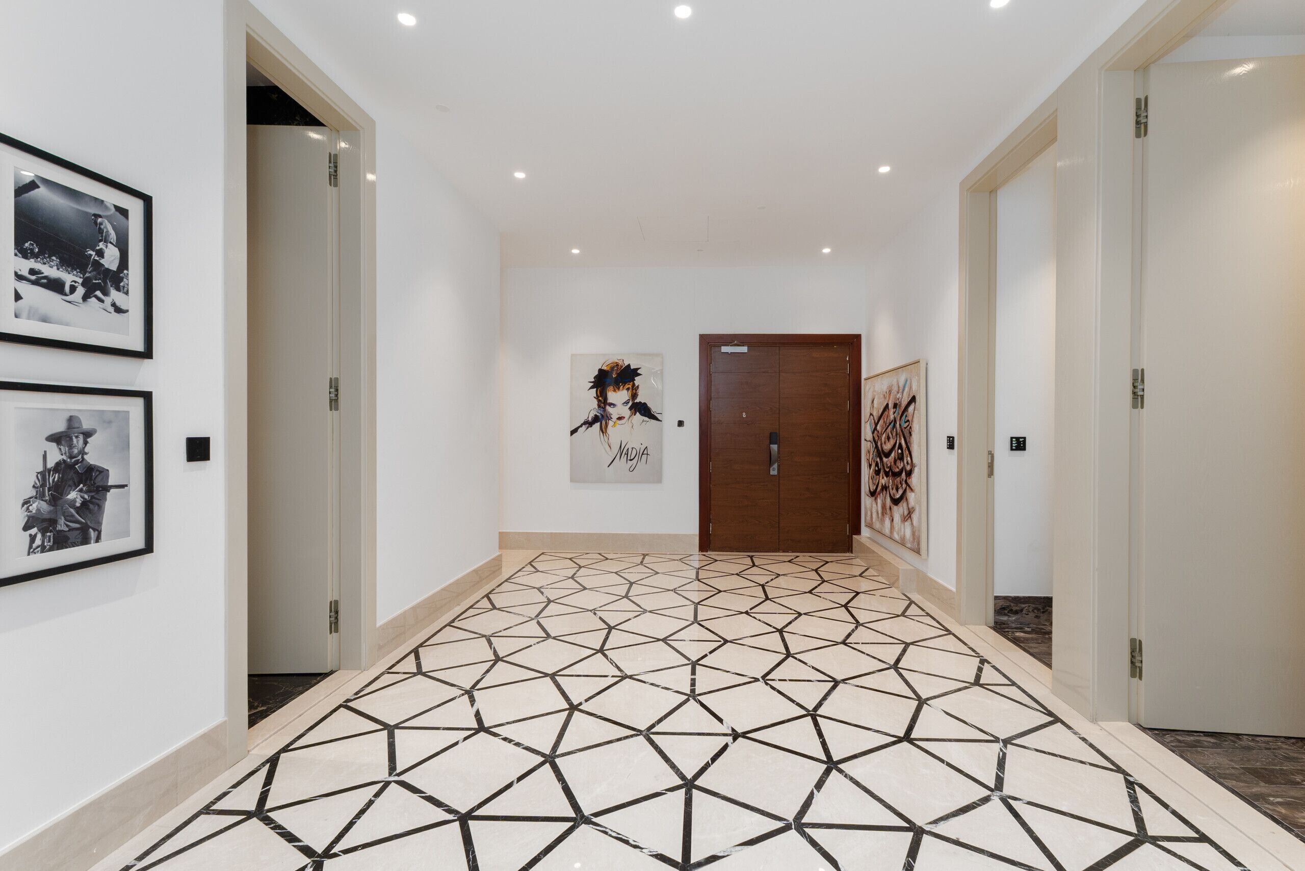 Ein Flur mit einem geometrisch gemusterten Boden, gerahmten Kunstwerken an den Wänden und einer Holztür am Ende.