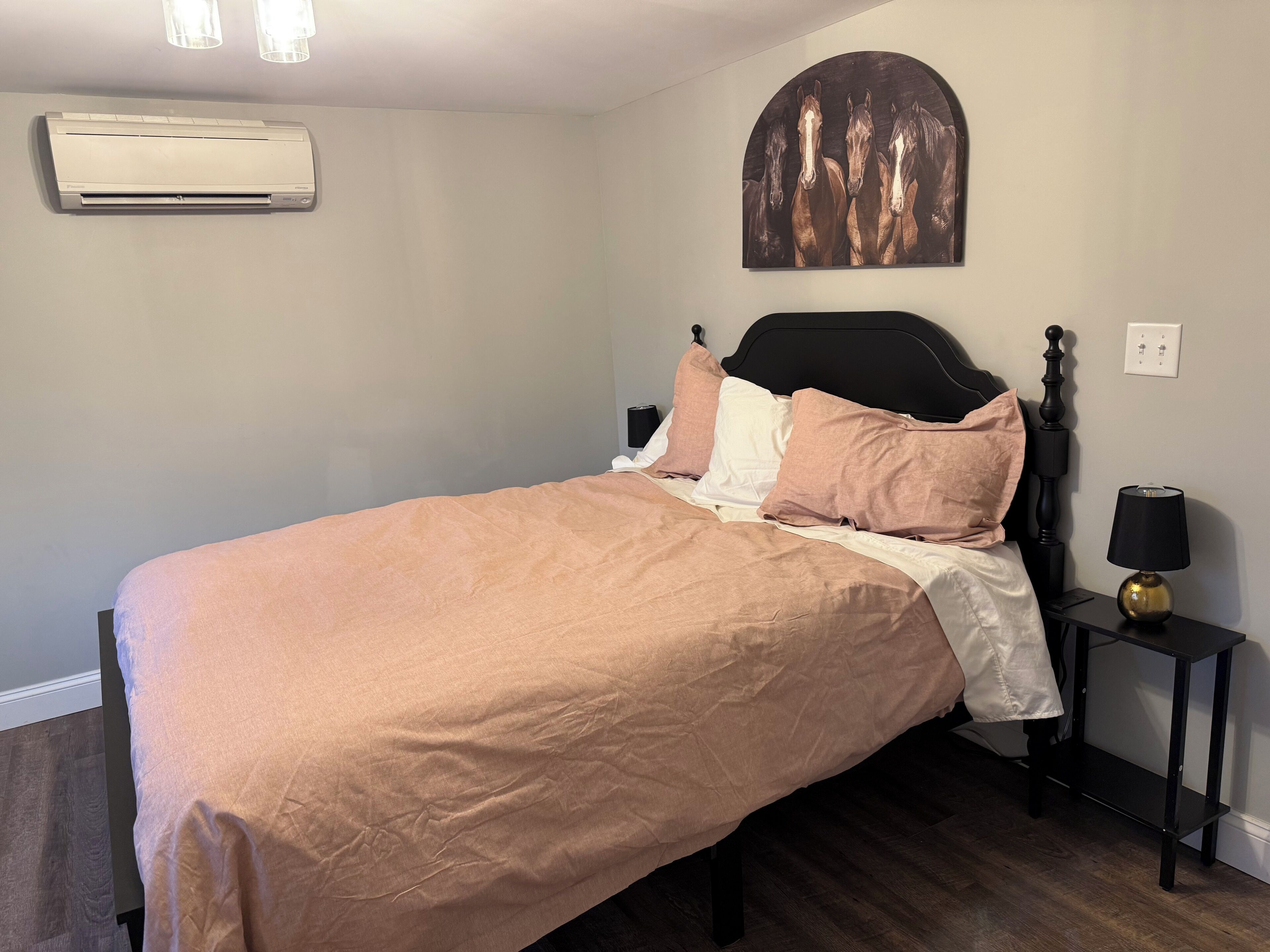 A bedroom with a bed, a wall-mounted air conditioner, a nightstand with a lamp, and a wall art piece.