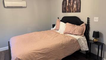 A bedroom with a bed, a wall-mounted air conditioner, a nightstand with a lamp, and a wall art piece.