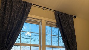 A bedroom with a large window, a bed with a beige quilt, and dark patterned curtains. - Circle F Ranch Bunkhouse- Charming farm stay in Shenandoah county- whole house (Edinburg)