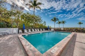A rectangular swimming pool with a tiled edge, surrounded by lounge chairs and umbrellas, with palm trees and a clear sky in the background.