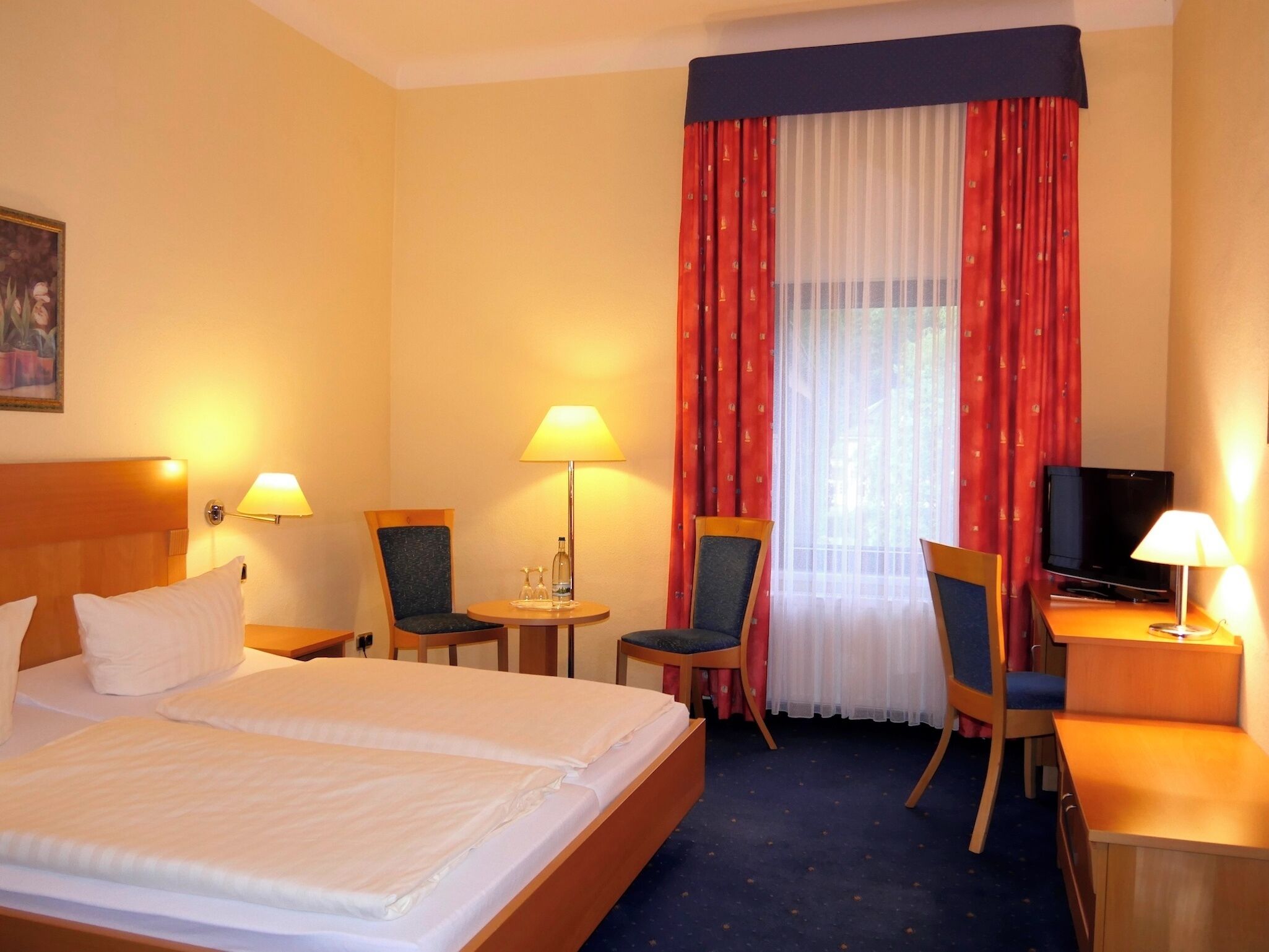 Une chambre d’hôtel avec un lit, deux chaises, un bureau avec une télévision et une fenêtre avec des rideaux.