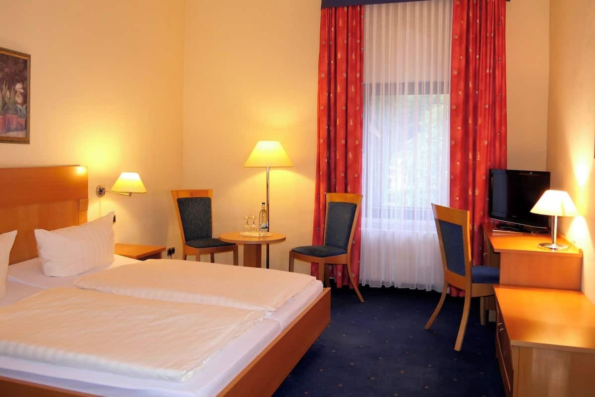 A hotel room with a bed, two chairs, a desk with a television, and a window with red curtains.