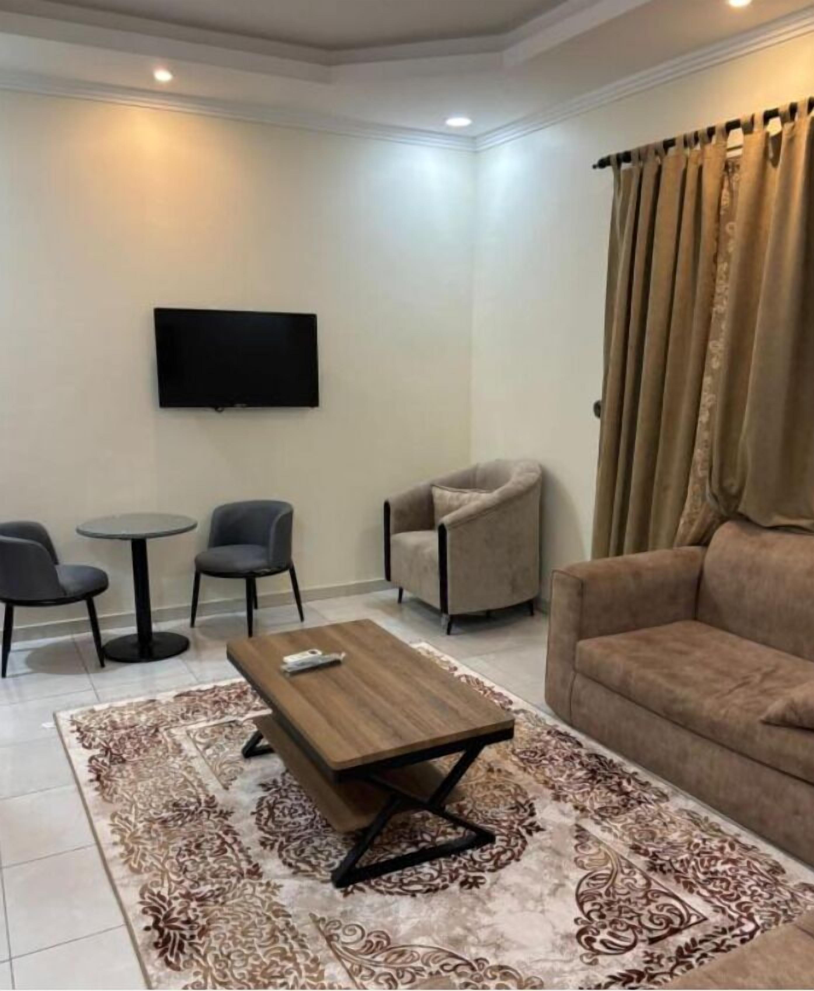 A living room with a brown sofa, a coffee table, two armchairs, a TV mounted on the wall, and a patterned rug.