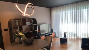Ett modernt vardagsrum med ett runt matbord, bekväma sittplatser, en inbyggd TV och ett stort fönster med tunna, genomskinliga gardiner.