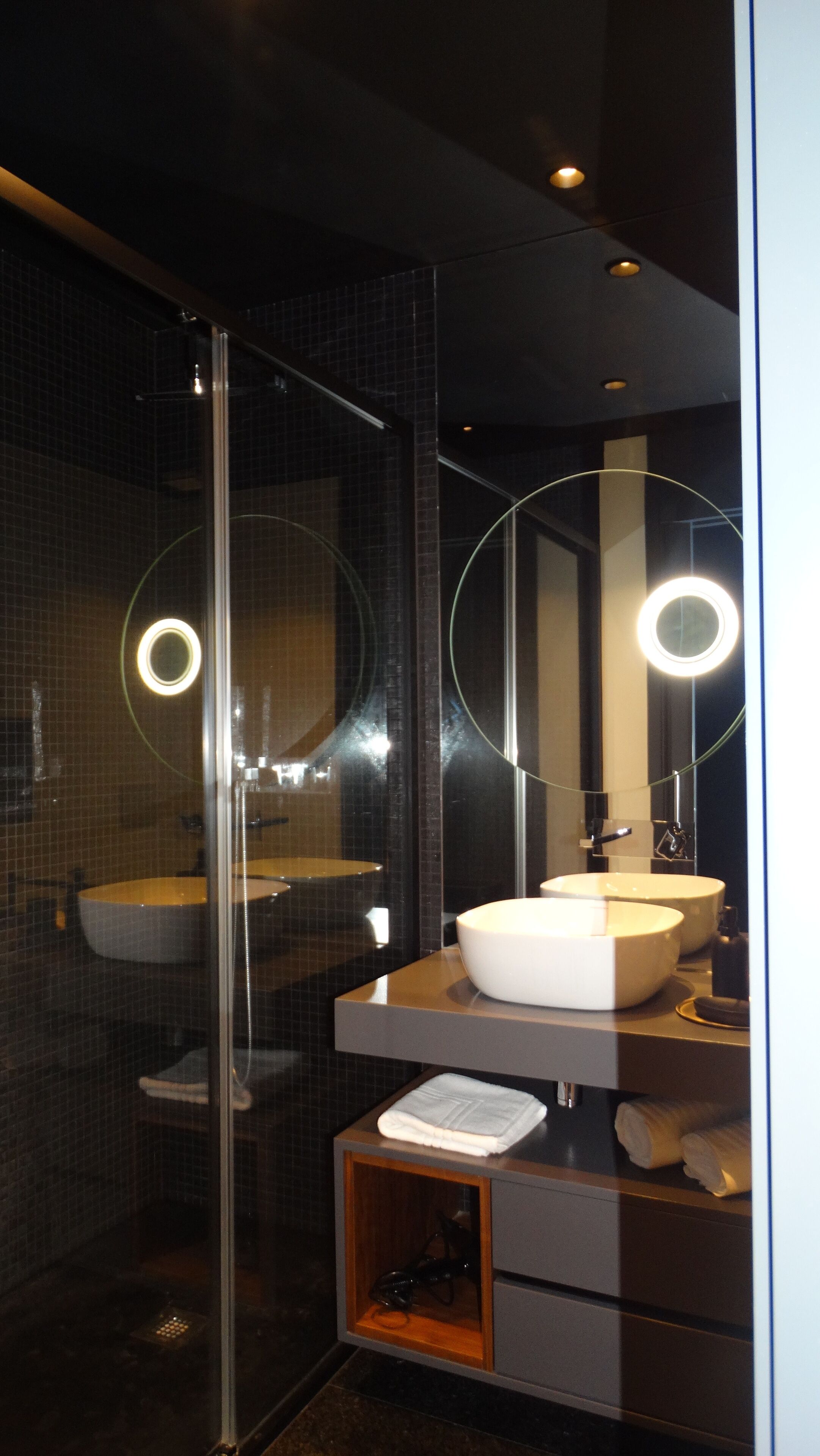 A modern bathroom with a glass shower enclosure, a white sink with a round mirror, and a wooden cabinet below.