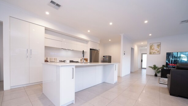 Una cocina moderna con armarios blancos y una isla central, una sala de estar con un sofá oscuro y un televisor de pantalla plana, y un amplio ventanal que proporciona luz natural.