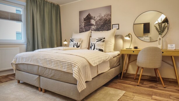 A neatly arranged bedroom with a bed, bedside table, lamp, chair, and a desk with a mirror. - Ankerplatz-Neumarkt - My vacation apartment in the heart of Bremerhaven! (Bremerhaven)