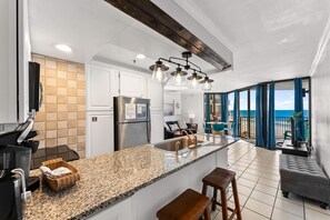 Eine Küche mit Granitarbeitsplatten, einem Edelstahlkühlschrank und einer Kaffeemaschine. Es gibt eine Sitzecke mit einer Couch und einem Tisch, sowie einen Balkon mit Blick auf den Ozean.