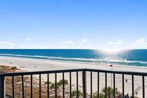 Blick auf einen Strand vom Balkon mit Geländer, feinem Sand, Meer und einer Flagge.