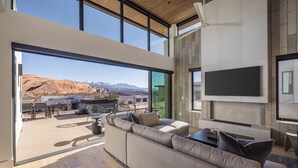 Una sala de estar moderna con un sofá grande, un televisor de pantalla plana montado en la pared y vistas a un paisaje montañoso a través de amplias ventanas.