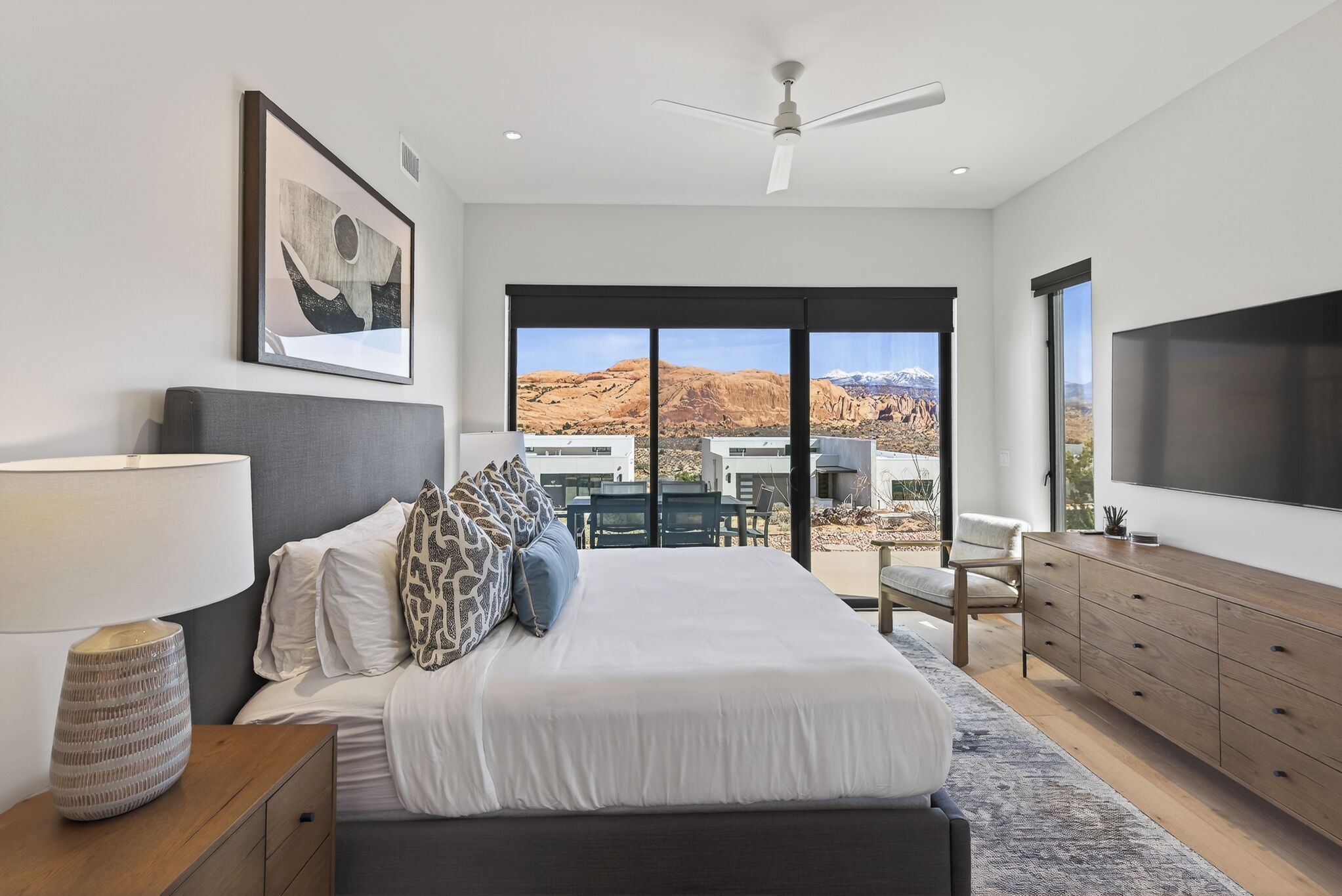 A bedroom with a bed, a TV, a ceiling fan, a large window with a view, and a wooden dresser.