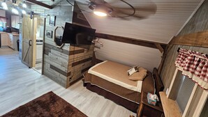 Une chambre confortable avec un lit, un ventilateur de plafond et un téléviseur fixé au mur, qui est en bois.