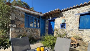 Un bâtiment en pierre avec une porte et des fenêtres bleues, une table sur laquelle se trouve une assiette de citrons, et des sièges à l’extérieur.