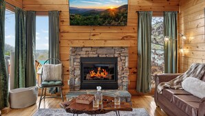 L’intérieur chaleureux d’un chalet avec une cheminée en pierre, un coin salon confortable et un téléviseur diffusant un paysage pittoresque.