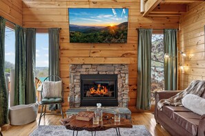 A cozy cabin interior with a stone fireplace, a comfortable seating area, and a television displaying a scenic view. - Smoky Mtn Views · Hot Tub · Game Room · Sleeps 12 (Sevierville)