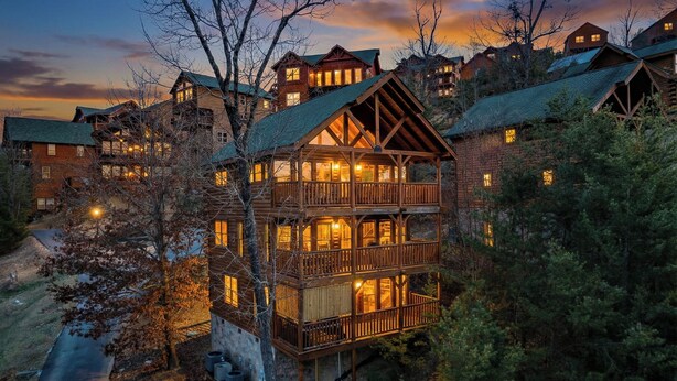 Une cabane en rondins à plusieurs étages, dotée d’un balcon, entourée d’arbres et se détachant sur un ciel crépusculaire.