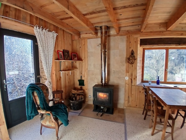 A wooden cabin interior with a wood-burning stove, a dining table, chairs, a chair with a blanket, a window, and a door.