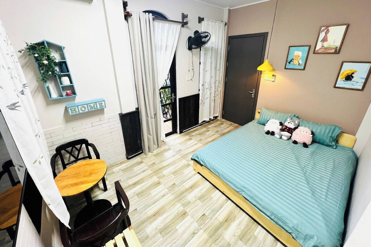 A bedroom with a bed, a desk, chairs, a small table, a fan, a lamp, and a door.