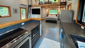 A modern kitchen with a built-in oven and microwave, a dining area with a wooden table, and a bedroom with a bed and a window overlooking greenery. - Modern Farm Cottage on Private Acreage in Charming Point Roberts, WA (Point Roberts)