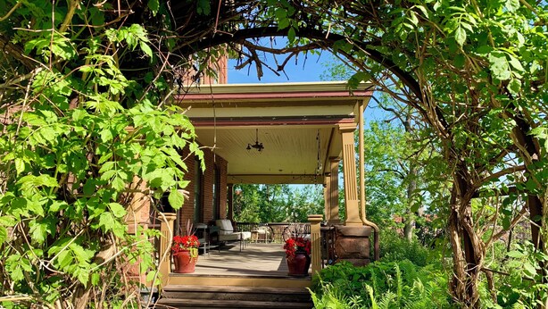 A covered porch with steps leading to a seating area, surrounded by greenery. - Quaint 7 bedroom mansion in charming Dubuque perfect for families (Dubuque)