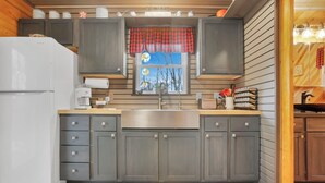 A compact kitchen with grey cabinets, a white refrigerator, a stainless steel sink, and a red and black checkered window valance.
