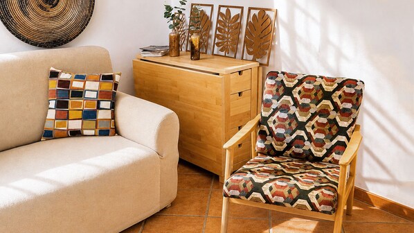 A beige sofa with a colorful pillow and a patterned armchair with a wooden side table in a room with a tiled floor and decorative wall art.