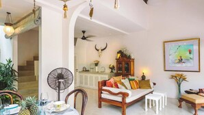 Ein großzügiger Wohnbereich mit Sofa, Esstisch und Stühlen. Ein Ventilator, Deckenventilator und eine Treppe in einen anderen Raum sind vorhanden.