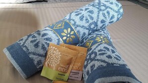 Un letto con una coperta a motivi blu e bianchi, due flaconi marroni di shampoo e balsamo ecologici e un piccolo cuscino blu e bianco.