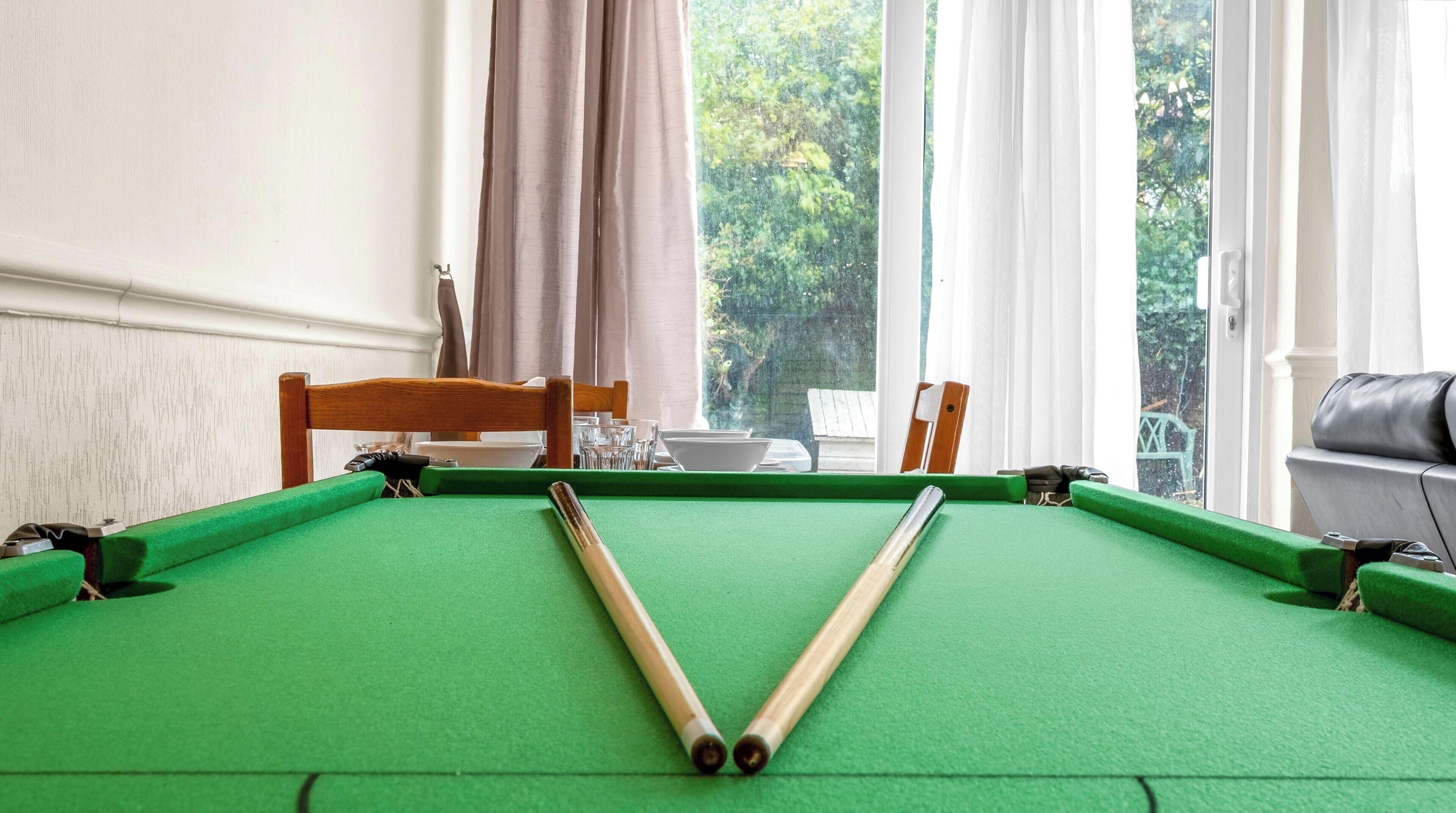 A pool table with a cue stick in the center, a wooden chair, and a view of a garden through a window.