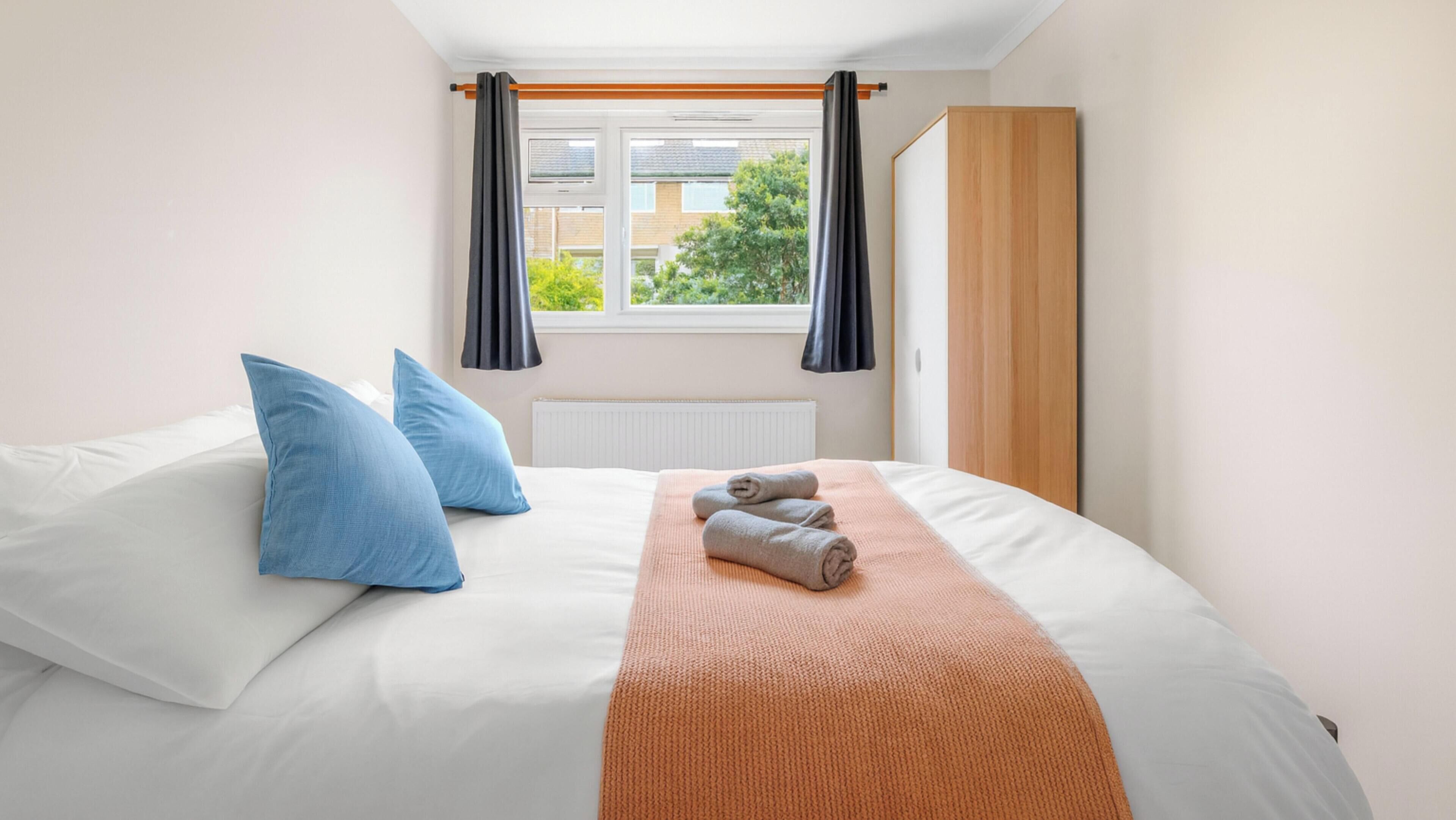 Een net opgemaakt bed met witte lakens, twee blauwe kussens en een opgevouwen handdoek.