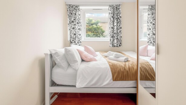Una cama bien tendida con ropa de cama blanca y rosa, un edredón beige y una toalla enrollada.